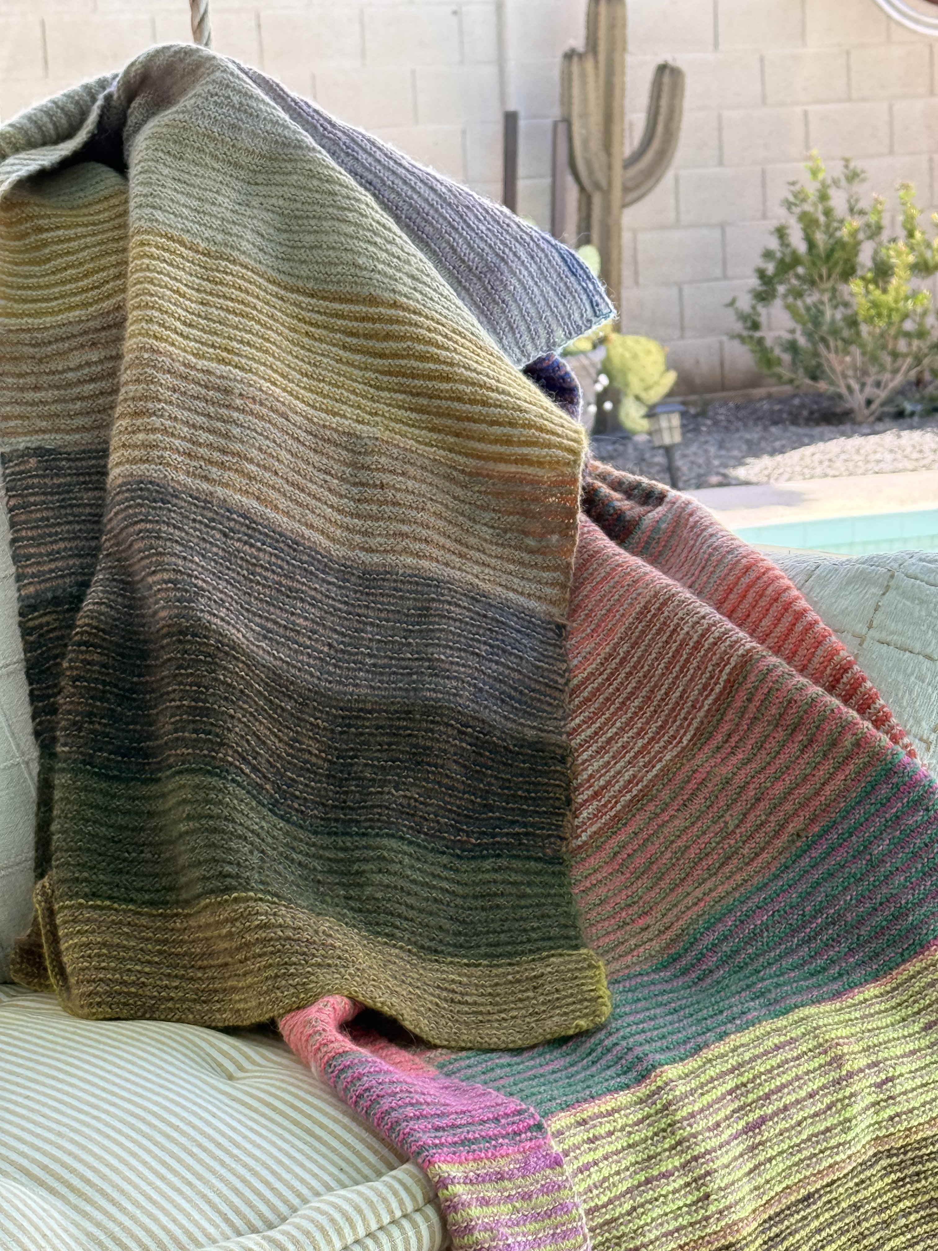 Alpenglow Blanket draped over an outdoor chair, showing the full spectrum of striped colorwork in Purl Soho Linen Quill Minis