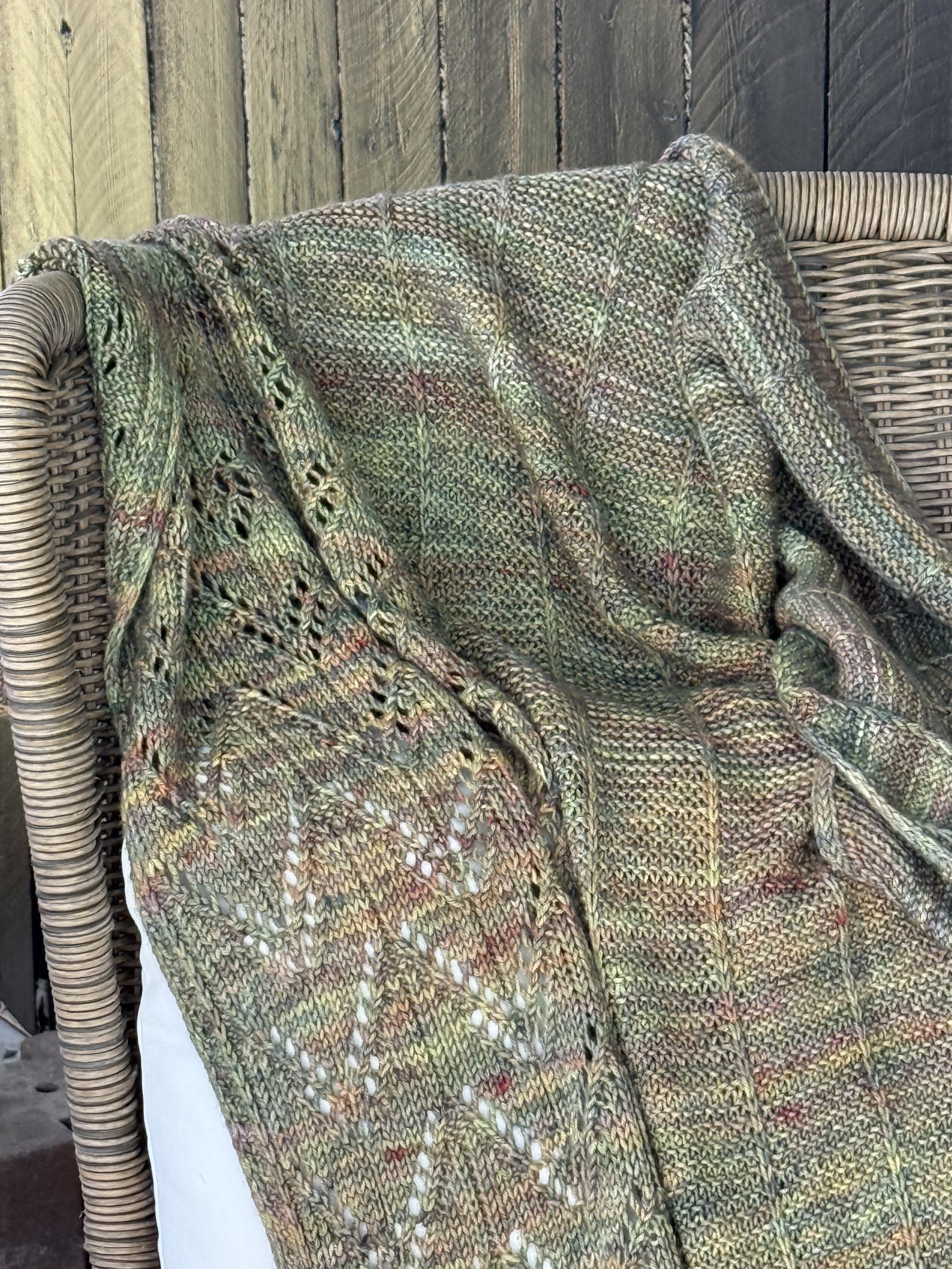 Kelcy's Garden shawl draped over a wicker chair, showing the full lace motif in sage green Malabrigo Rios