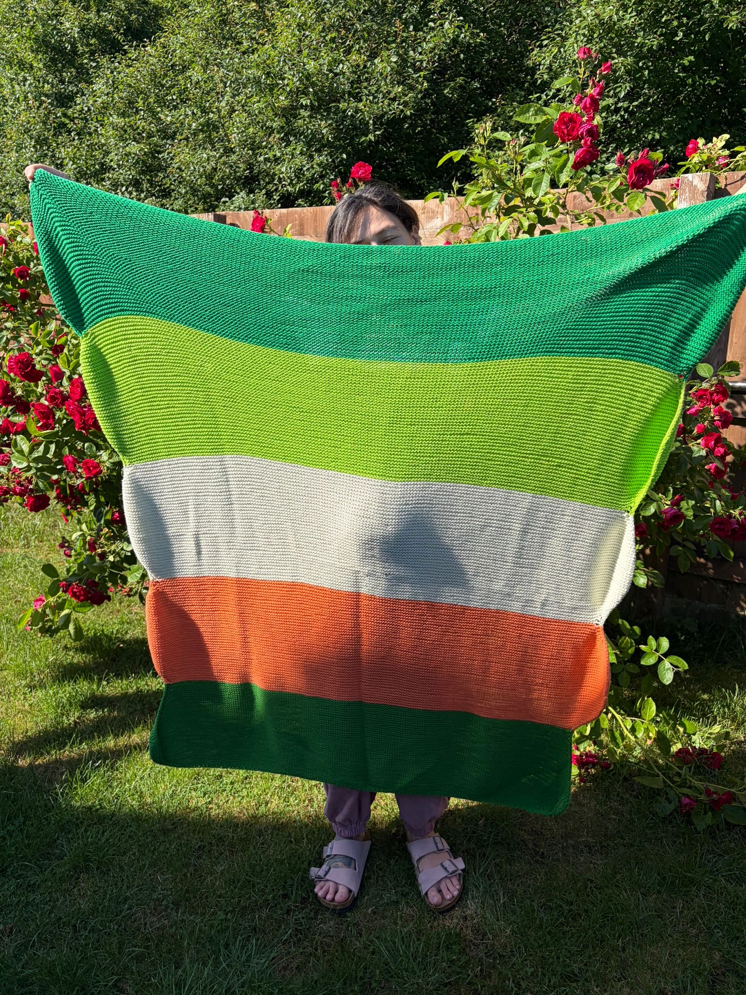 Macaron Blanket in Carrot Top held up outdoors, showing bold horizontal stripes in emerald, lime, cream, and terracotta against a backdrop of red roses