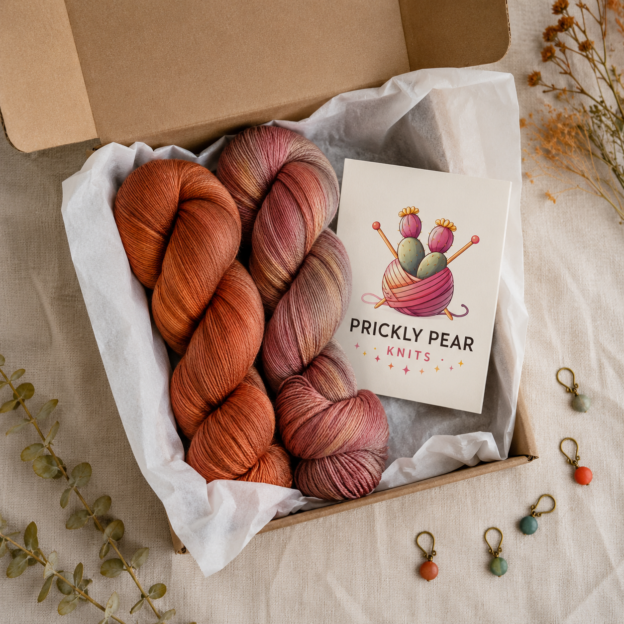 An open Prickly Pear Knits subscription box with two hand-dyed yarn skeins, a branded card, and stitch markers